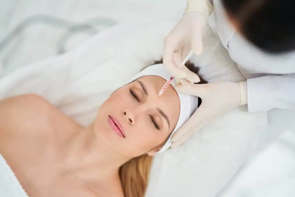 A woman lying down with her eyes closed while a medical professional injects a cosmetic treatment into her forehead.
